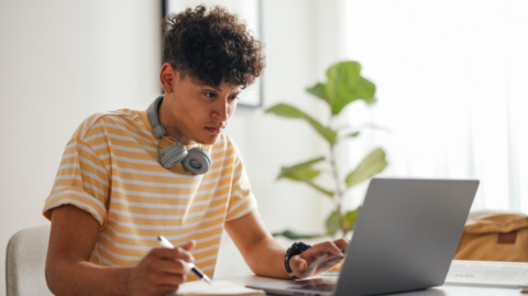 A male in his late teens or early 20s deep in work mode at a tidy, sunlit desk. They’re wearing a yellow‑and‑white striped shirt with grey headphones resting around their neck, one hand on a pen as they write in an open notebook beside their laptop. A leafy green plant sits in the background near a bright window