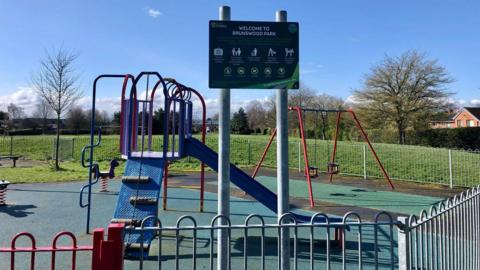 A play area - at the front of the photograph is a sign saying "Welcome to Brunswood Park" - behind it is a blue slide. Towards the back of the park is a play horse on a spring and a set of swings.  The park has railings around it.