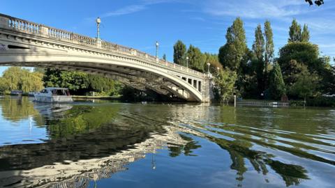 A bridge over a river stands in glorious sunshine with a boat sailing underneath it. There are trees lining the river.