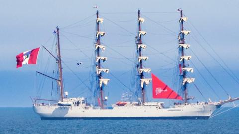 A tall ship. Four towering masts fully dressed in canvas. On the left side, the bold red-and-white flag of Peru flutters as well as a red triangular sail on the right bearing the word "Peru" and a swirling spiral logo. The sky behind is blue and visibility is clear.