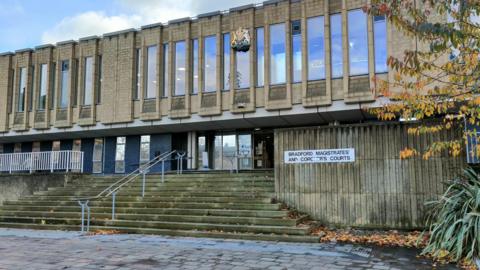 A 1960s court building in light stone. It has high narrow windows. On the right is a sign that reads: 'Bradford Magistrates' and Coroner's Courts'. There are some fallen orange leaves on the street.