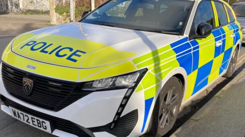 A Devon and Cornwall Police car parked on a residential street. It is white, yellow and blue. The make of the car is Peugeot.