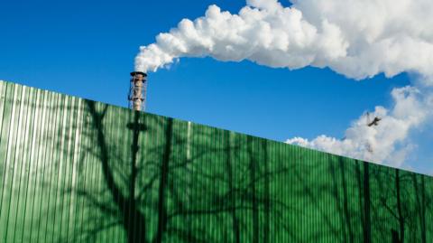 A stock image of chimneys with vapour coming out of the top, behind a green industrial fence.