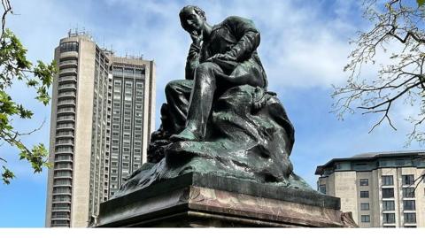 A bronze statue on a marble plinth, surrounded by the tall buildings on Park Lane. The statue depicts Lord Byron seated, with his head resting on his right hand in a pensive mood. 