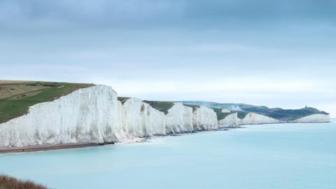 The white cliffs at Cuckmere Haven and Beachy Head surrounded by a calm sea. In the foreground two walkers stroll along the grass on the clifftop and overlook the view. 