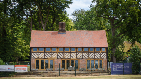 Restored historic timber-framed building with a red tiled roof and brick ground floor, viewed from across a grassy field. Large glass doors line the front façade, with mature trees surrounding the property and conservation project signs displayed along the perimeter fence.
