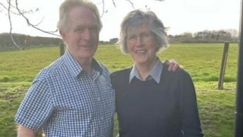 Campbell Welsh dressed in a blue and while gingham short-sleeved shirt with his arm around his wife Sheila, who is wearing a shirt with a collar underneath a navy pullover. They are standing in a field.