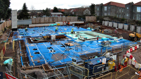 The site as it was on 2 December 2024 with a big blue tarpaulin material across the base of the site and various tubes exposed with diggers, and exposed soil around the outer edges. There are also people on the site in high vis and hard hats.