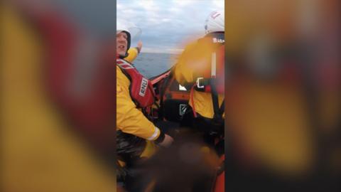 Two RNLI rescuers - dressed in lifejackets and yellow uniforms - are on a lifeboat in the sea. One points ahead