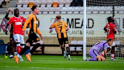 James Brophy celebrates after scoring for Cambridge United, running away from goal pointing towards teammate Elias Kachunga, who set up the goal, as Charlton Athletic players look dejected