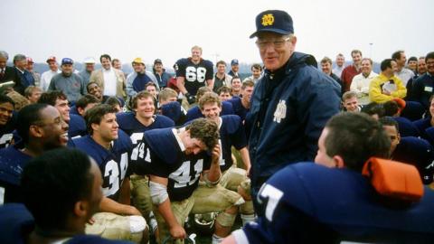 Lou Holtz standing at the centre of a group of kneeling players, and speaking. He's in a baseball cap and windbreaker with the Notre Dame logo 