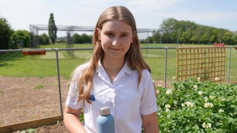 A teenage girl stands outside in a school sports field. She is wearing a white school shirt and holding a blue water bottle. She has long blonde hair and is smiling.