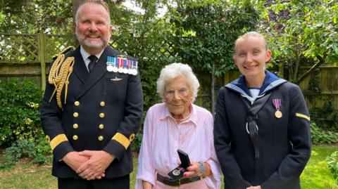 Mrs Farmers stands in a garden holding an open box containing a medal, flanked by two individuals in formal military uniforms. Green foliage and a wooden fence are visible in the background."