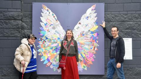 Three people stand around the artwork made of feathers that are placed in a way that makes them look like a pair of wings. One woman stands in the middle, as if they are her wings, and is smiling at the camera. A man and woman stand on either side of the piece, also smiling at the camera.