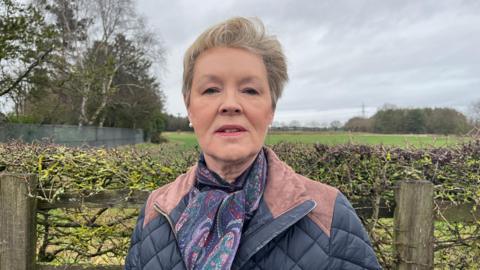 Susan pictured looking into the camera with a neutral expression. She has short blonde hair, and is wearing a navy blue quilted jacket with light brown suede patches on the collar and shoulders. She's also wearing a dark blue scarf with a purple, red and green paisley pattern on it. Susan is stood in front of a hedge, which is bracketed by a wooden fence. Behind her, a grassy field stretches into the background, with a row of trees running along the left side of the photo. In the far distance, and slightly out of focus, electricity pylons can be seen. The sky is grey and overcast. 