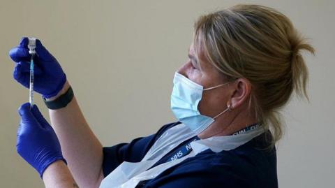 A nurse preparing a vaccine. She is wearing a blue uniform and NHS lanyard and she has blue plastic gloves, a white plastic apron and a mask. Her hair is tied back.