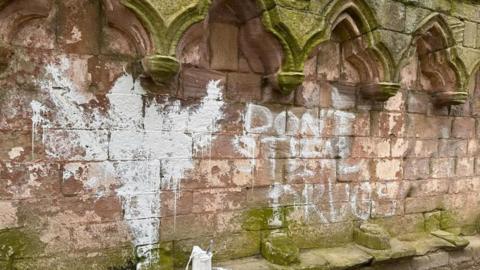 The words "DON'T STEAL DRUGS" have been daubed in white paint on the wall of a medieval abbey. There is also a large splodge of paint on the wall to the left. A can of white paint has been left nearby. 