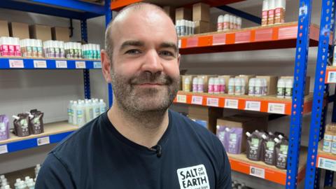 Man in blue "salt of the earth" t-shirt stands in front of shelves with bottles and pouches on them