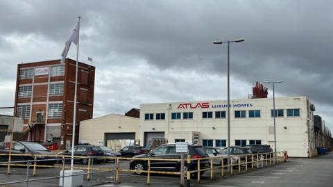 The exterior of Atlas Leisure Homes' headquarters on Wiltshire Road in Hull. The main building is covered in cream cladding and has a large blue and white logo spanning the width of the building. A building on the left of the image is a red brick tower block with six large windows. A car park is in front of the two buildings along with a flag pole and lights.