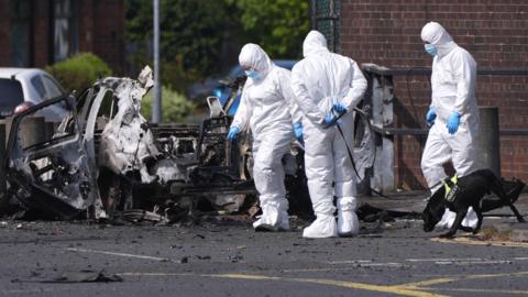 Three forensic investigators dressed in white hazmat suits, blue plastic gloves and blue medical masks walk in front of the wreckage of a car bomb, examining the ground. The vehicle has been blown apart. One of the investigators is holding a dog lead which is tethered to a slim black Labrador. The dog is wearing a bright yellow harness which says: "Police K-9". 