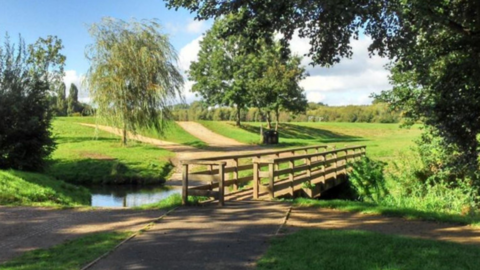 A wooden bridge over a waterway, it is sited in a park with trees behind it and a pathway on either side