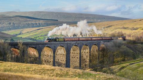 The West Yorkshireman service, hauled by a steam locomotive, travels on the Settle to Carlisle line over the Ribblehead Viaduct. The charter is pulling several brown carriages. The views span over the Yorkshire Dales National Park and the Pennines. 