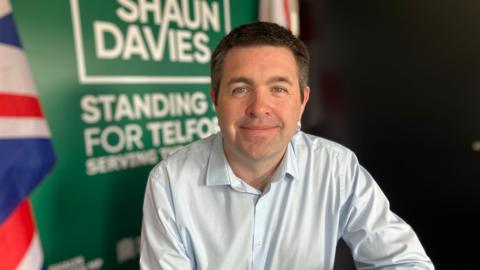 Shaun Davies has short dark brown hair and is sitting down, wearing a light blue shirt. He has a green sign behind him that says "Shaun Davies Standing for Telford".
