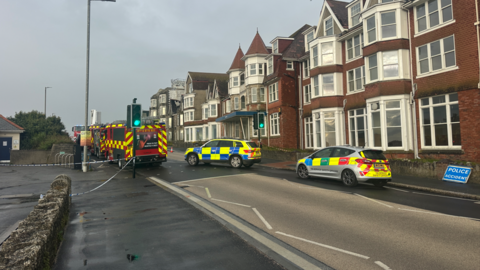 A picture of the road where the fire took place. It is a large road with emergency vehicles present on scene. A number of houses can be seen to the right of the road.