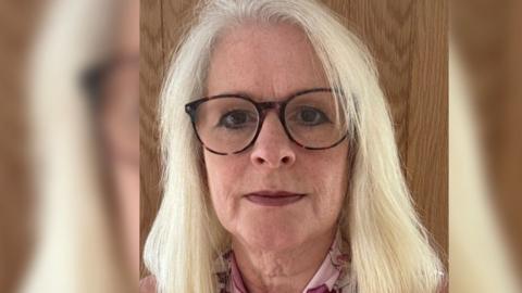 Close up of Sue's face, she has blonde hair and black glasses with brown eyes. She is stood in front of a wooden frame.