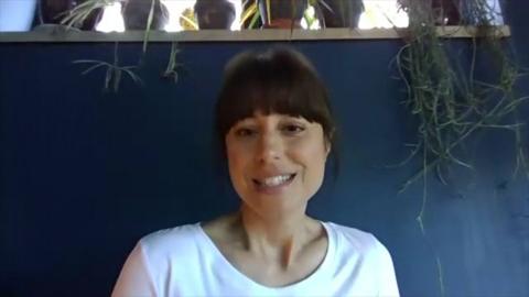 Zahra Ahmadi looking at the camera and smiling. She is sat against a blue wall with a hanging plant on the right.