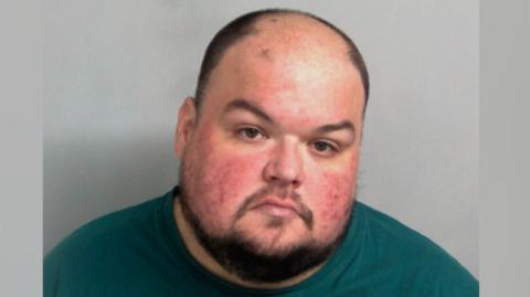 Gavin Plumb looks serious, if not slightly angry, in a picture taken in police custody. He has a chinstrap beard and a moustache. He is wearing a green T-shirt.