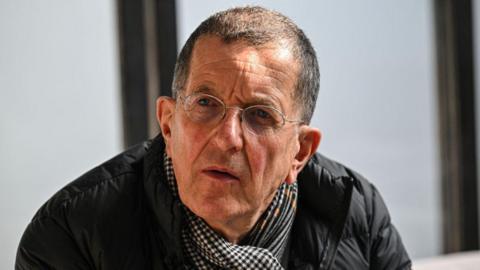 A close-up image of Sir Antony Gormley. He has short dark hair and is wearing wire-rimmed glasses, a black jacket and a checked scarf.
