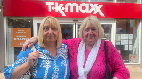 A woman with blonde hair is on the right wearing a pink top and has her right arm around another woman with blonde hair on the left, who is wearing a blue and white top. They are in front of TK Maxx.