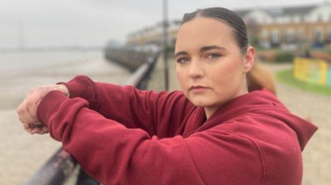 Danielle wearing a red hoodie looking to camera leaning over a railings on an unidentified seafront