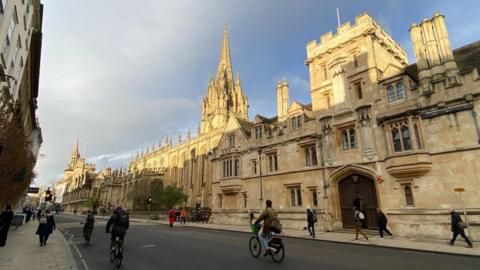 Cyclists and pedestrians are seen meandering along a road in Oxford in front of the city's famous spires