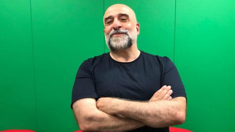 Comedian Omid Djalili is wearing a black T shirt and has his arms folded. He stands in front of a grey background with a slight smile