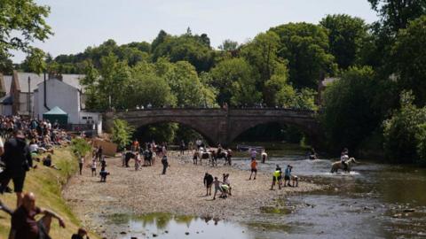 View downriver of people attending the Appleby Horse Fair. People are lining the banks of a river to watch riders taking their animals into the river. Others are watching from a bridge. The weather is sunny.