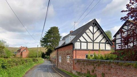 A cottage stands near a road with a walled garden. The walls are partly brick and partly black and white. Across the road, there is a grassy verge and hedge. Further cottages are around the property. There is open countryside further away.
