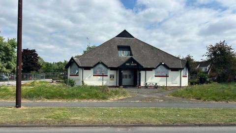 A pub with a large grey triangular roof with grass and a path in front of it.