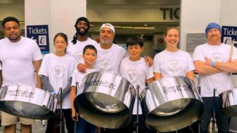 Eight people from the steel band. they are in white t-shirts that say "impact steel band". They are standing in front of three silver steel drums.