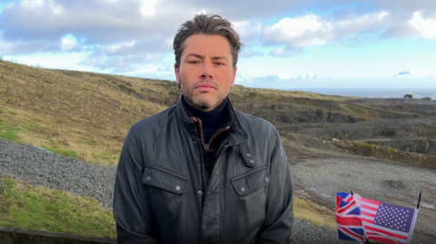 A man with grey and brown hair and grey facial hair is standing on a hill with a quarry in the background. There is green grass and hills behind him. To his right are two small flags of Great Britain and the USA