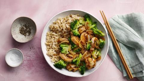 A white bowl with brown rice, chicken and broccoli in it. Beside the bowl are a pair of wooden chopsticks
