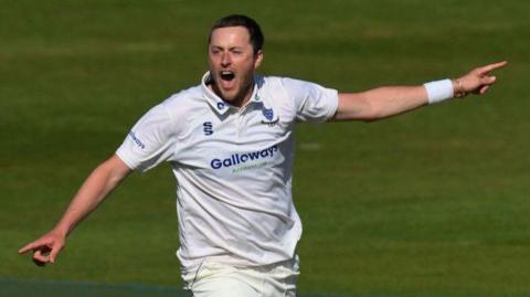 Sussex bowler Ollie Robinson celebrates a wicket with both arms out wide pointing, shouting, wearing club whites and sussex crest, with white wrist band on left wrist partially coveri ng siver bracelet