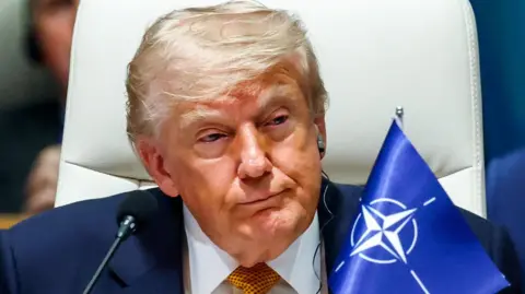 Trump at a meeting during the Nato summit. There's a blue flag next to him and he's wearing an earpiece.