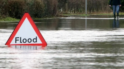 A river of floodwater swamping a rural road. In the foreground, a red and white triangular warning sign says "Flood". In the distance, someone wearing rubber boots is wading through the water.