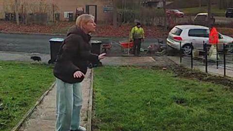 Woman arguing with neighbour in her front garden with two workmen in high-vis jackets standing behind her.