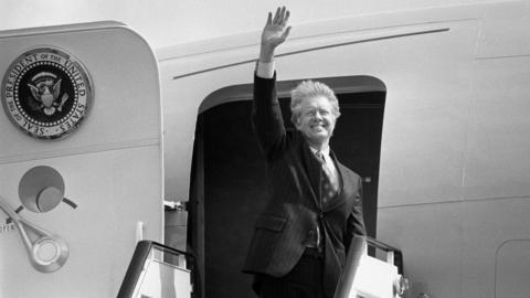 Jimmy Carter waving at the door of Air Force One which has the president's seal on the door - a circular badge with an eagle in the centre. Carter has grey, windswept hair.