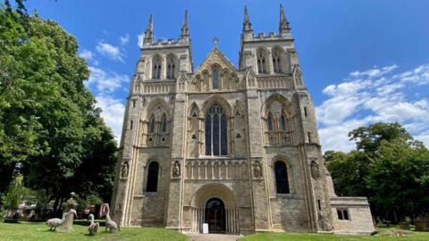 A picture of a place of worship made of sand-coloured limestone. The front of the building consists of a large, arching doorway and two towers to either side. Four statues feature next to the first two first floor windows.