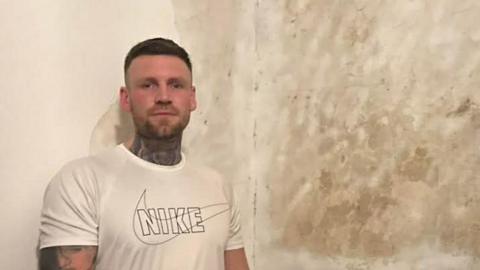 Josh Gallagher, wearing a white Nike t-shirt, stands pointing at the wall covered in black mould and damp.