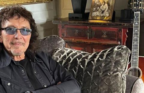 A man wearing tinted spectacles and a dark jacket sits on a sofa, with a guitar propped up against it